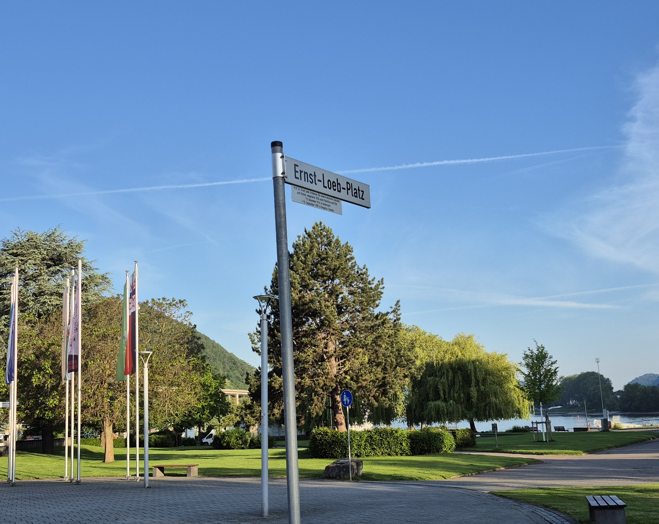 Ein Straßenschild mit der Aufschrift "Ernst-Loeb-Platz" steht in einem Park an einem Fluss